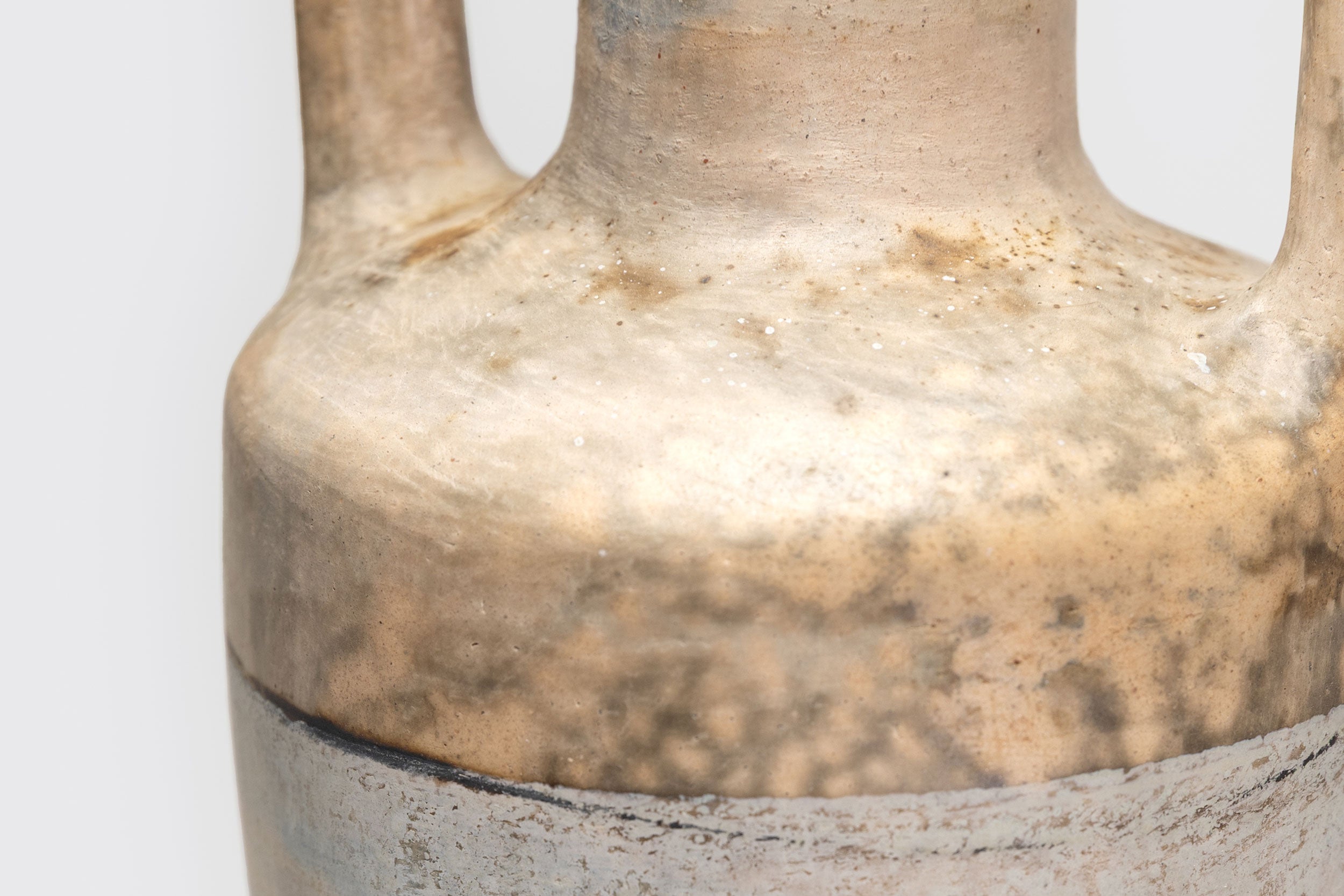 Close-up da parte superior de Ânfora#1, de Derek Tasker-Norton, um vaso de cerâmica artesanal envelhecida com duas alças e texturas manchadas de marrom, bege e cinza em sua superfície. Fundo claro e liso.