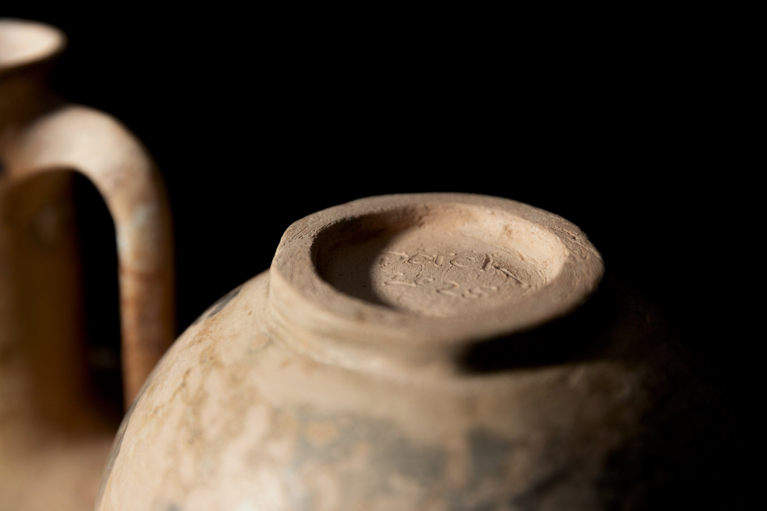 Close-up da base da Ânfora#3 de Derek Tasker-Norton, um pote de barro desgastado com marcações gravadas, ao lado de outro vaso de cerâmica artesanal, ambos colocados contra um fundo escuro.