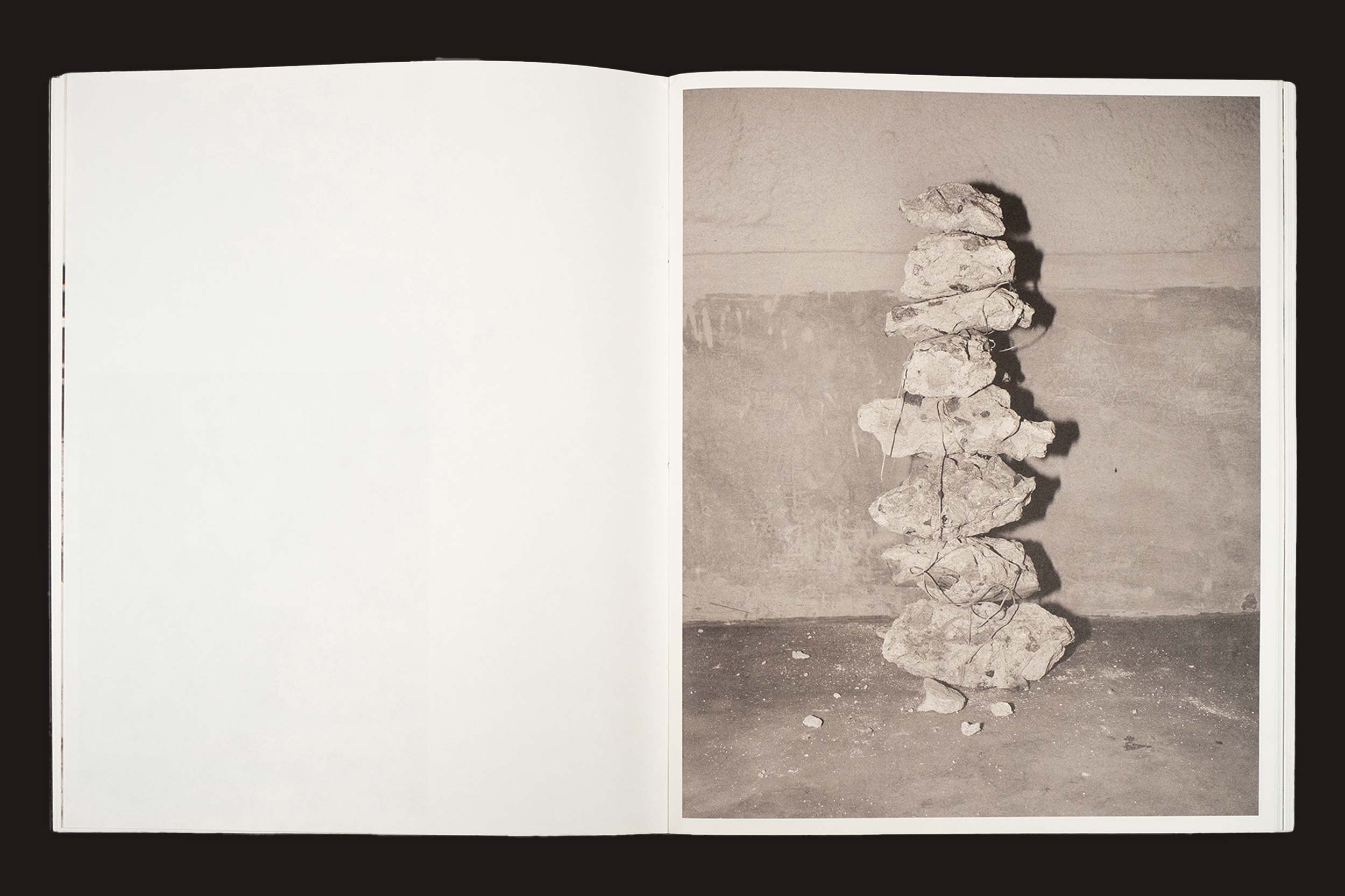 Uma página aberta de Bharat Sikka: Ripples in the Pond (Fw:Books) revela uma foto em preto e branco de pedras empilhadas e amarradas com arame formando uma escultura vertical contra uma parede áspera, com a página esquerda em branco.