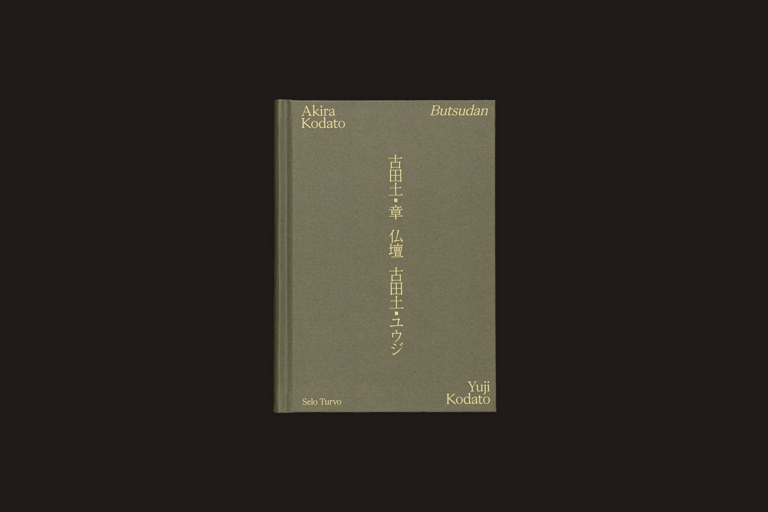 Um livro bilíngue de capa dura verde, "Akira Kodato & Yuji Kodato: Butsudan", da Selo Turvo, apresenta textos em inglês e japonês dourados na capa, com os nomes dos autores nos cantos e Sola Turato no canto inferior esquerdo, evocando memórias e heranças.