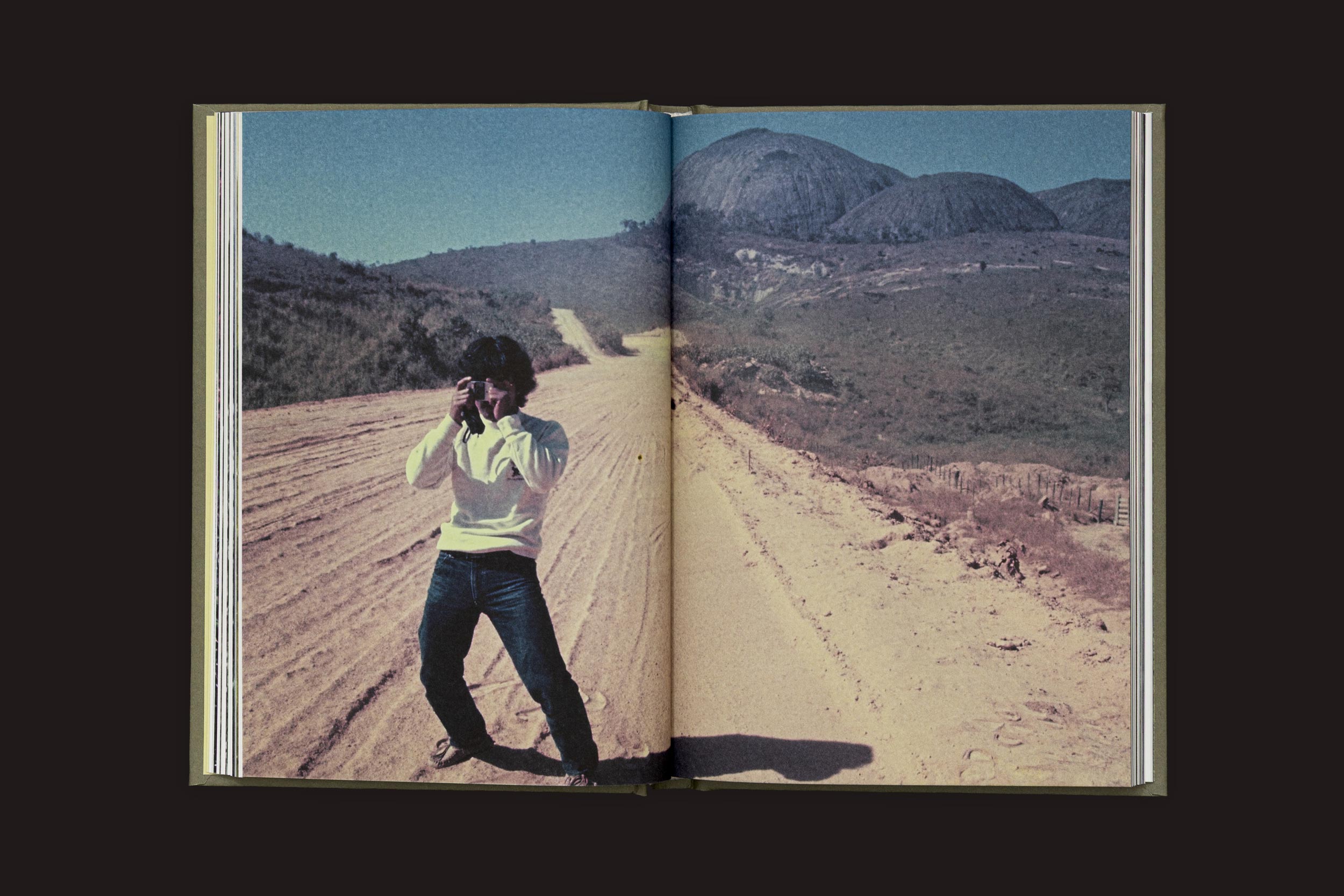 Um livro aberto de "Akira Kodato & Yuji Kodato: Butsudan", de Selo Turvo, mostra uma pessoa em uma estrada rural empoeirada entre a flora da montanha, com céu limpo e campos de café distantes, capturando memórias de herança ancestral.