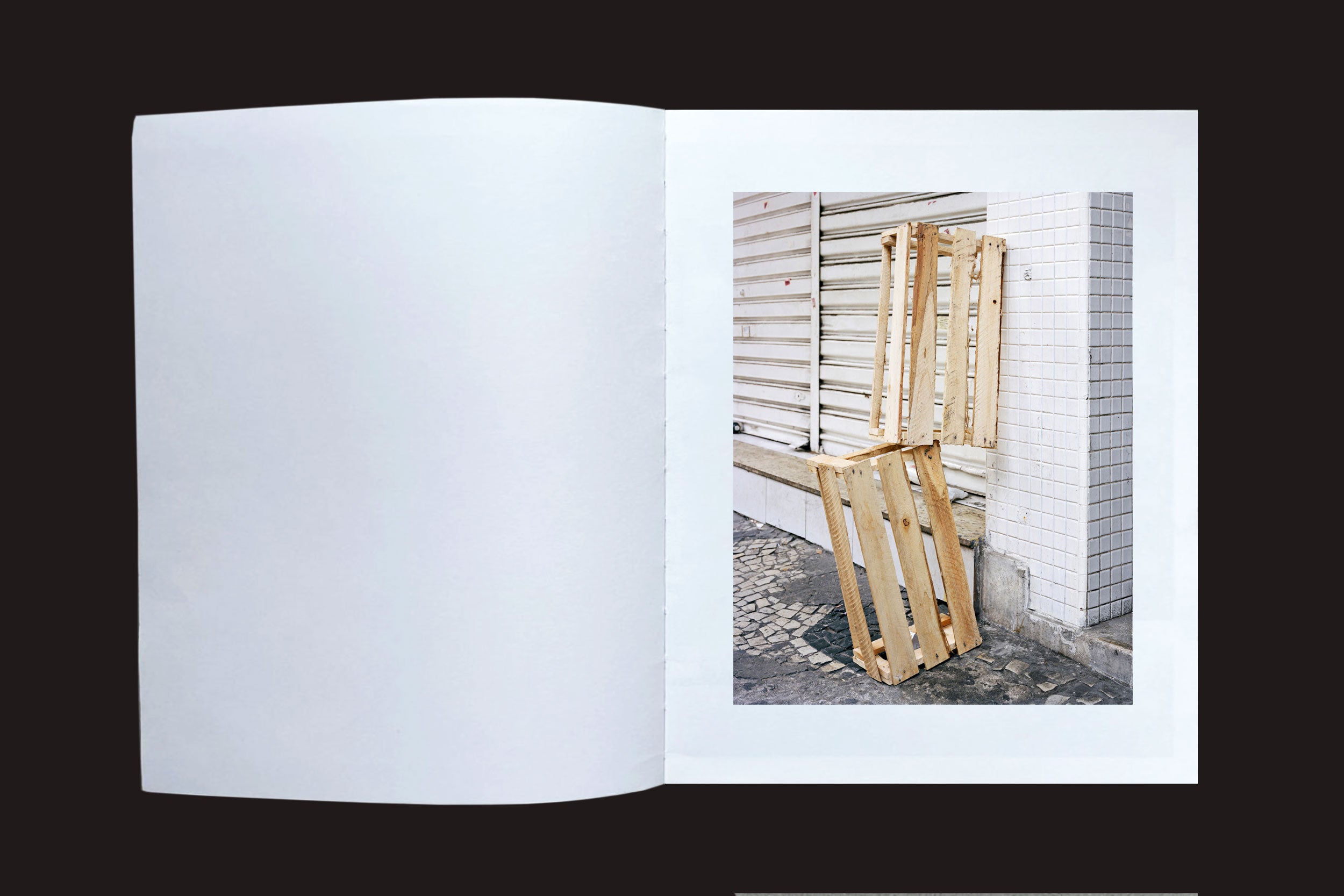 Em "Felipe Russo: Centro" da Auto-edição, um livro aberto mostra uma fotografia urbana: dois paletes de madeira contra uma parede de azulejos em uma calçada de paralelepípedos no centro de São Paulo, com a página esquerda em branco.
