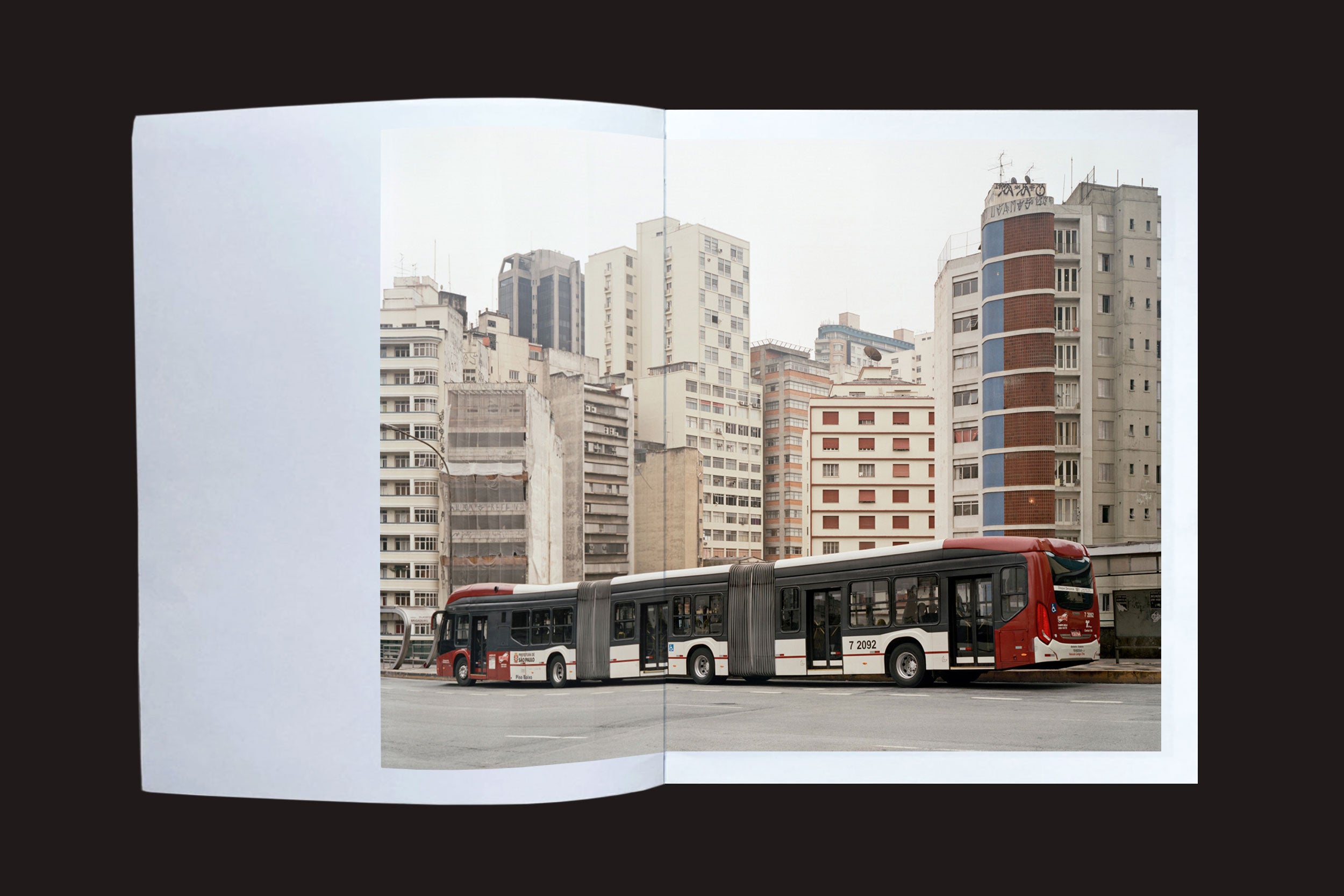 Uma foto de "Felipe Russo: Centro" da Auto-edição, mostrando dois ônibus urbanos articulados em frente a apartamentos modernos, é exibida em um livro aberto contra um fundo preto.