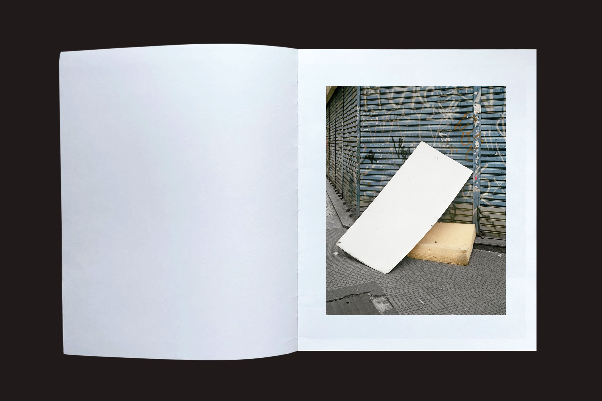 Um livro aberto da exposição "Felipe Russo: Centro" exibe uma fotografia urbana de um quadro branco ao lado de uma caixa de madeira diante de um obturador coberto de grafite, capturando a essência da paisagem urbana de São Paulo.