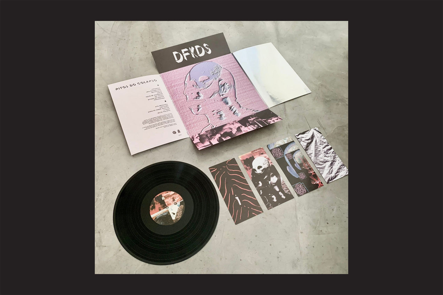 Um vinil de Deaf Kids: Ritos Do Colapso, da Nada Nada Discos, é exibido em uma superfície cinza com seu rótulo impresso, ao lado da capa do álbum desdobrada, da folha de letras e de três cartões ilustrados que mostram seu estilo punk transcendental brasileiro.