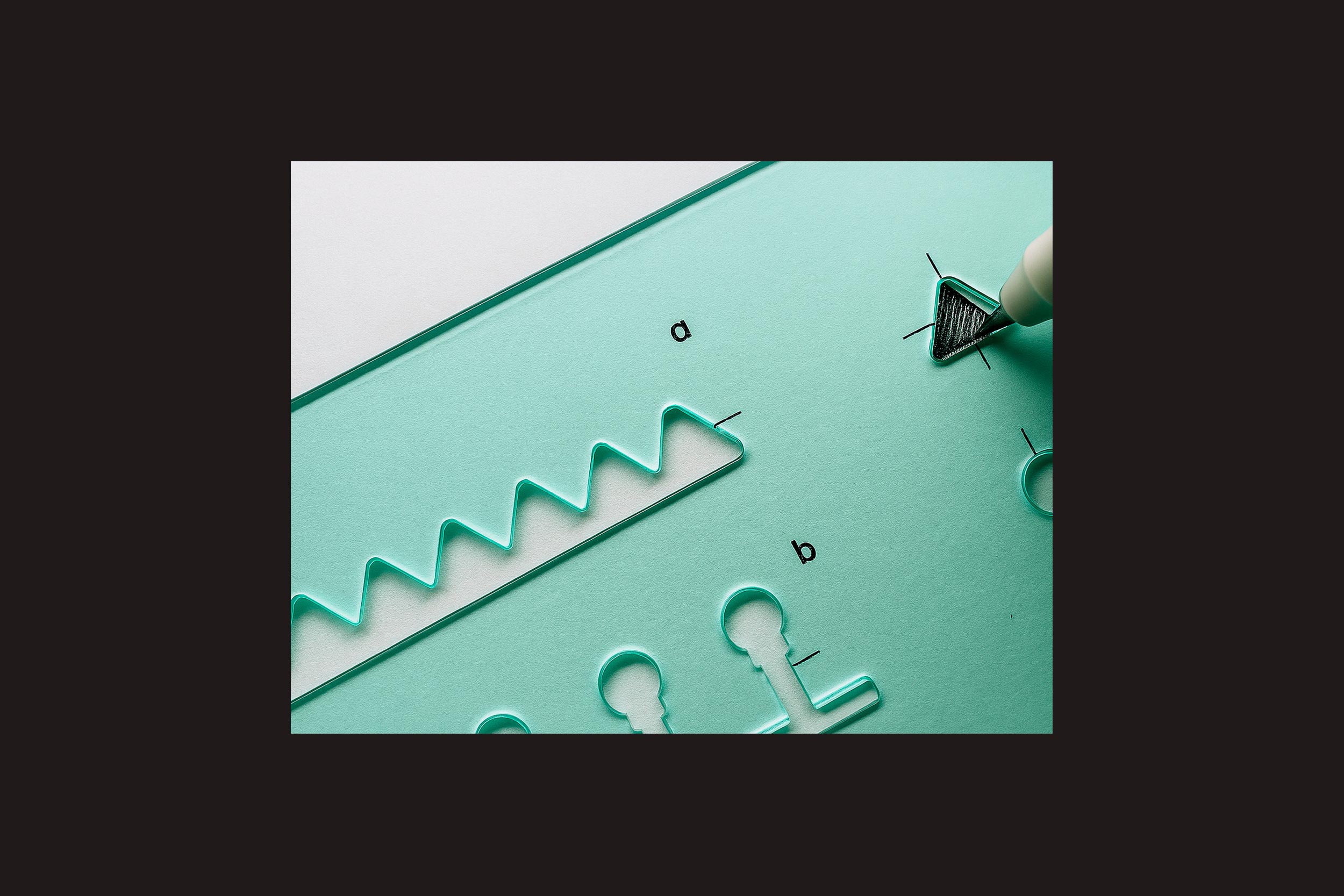 Um close-up de um estêncil Ermãos Monjon: Gabarito em papel branco apresenta formas geométricas e linhas onduladas rotuladas como "a" e "b". Uma mão usa uma caneta para traçar um triângulo, ilustrando essa ferramenta da Ermãos Monjon para arquitetura urbana hostil.