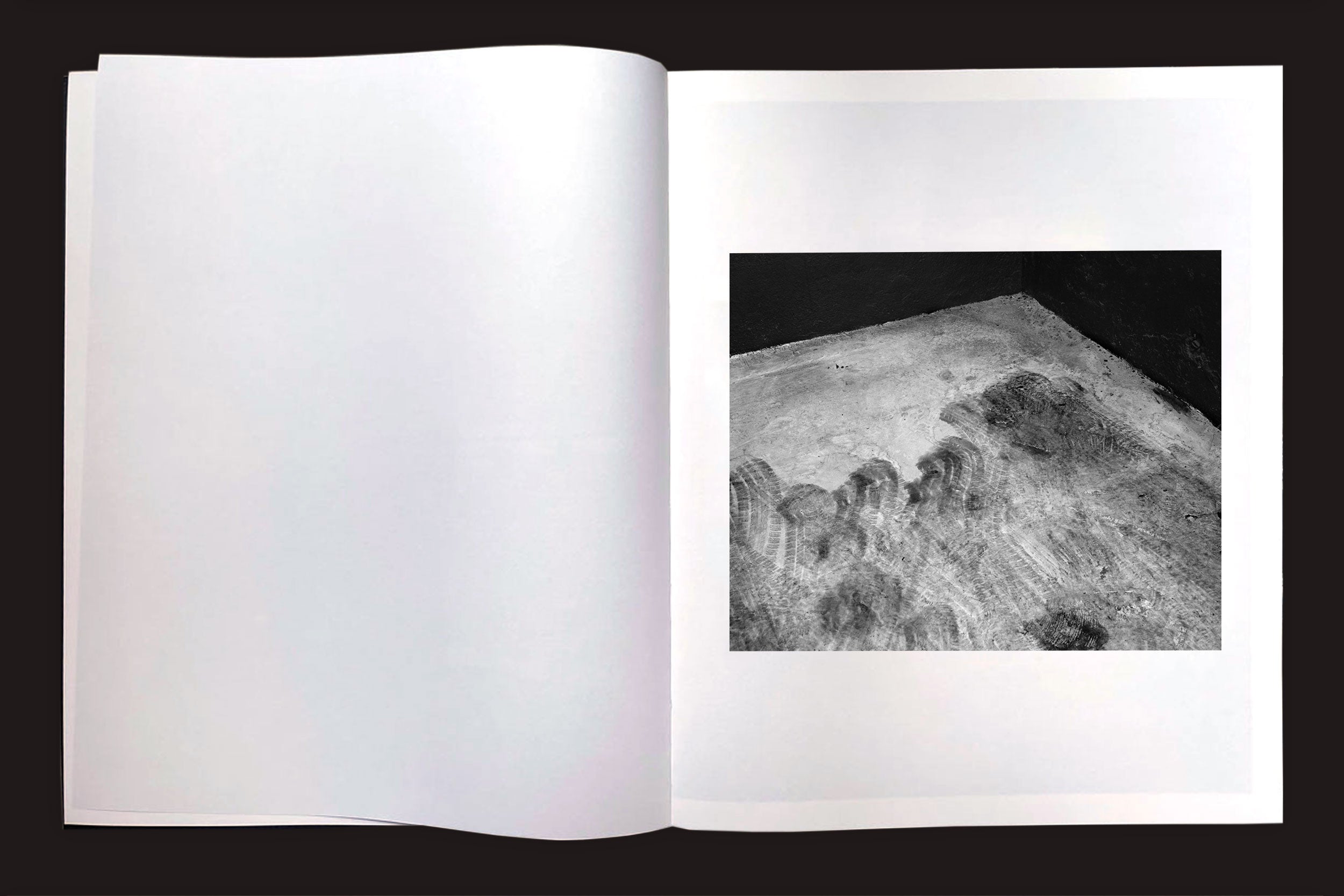 Um livro aberto, "Felipe Russo: Garagem Automática" da Auto-edição, exibe uma fotografia arquitetônica em preto e branco de um piso de concreto texturizado e sombrio na página direita; a página esquerda está em branco.
