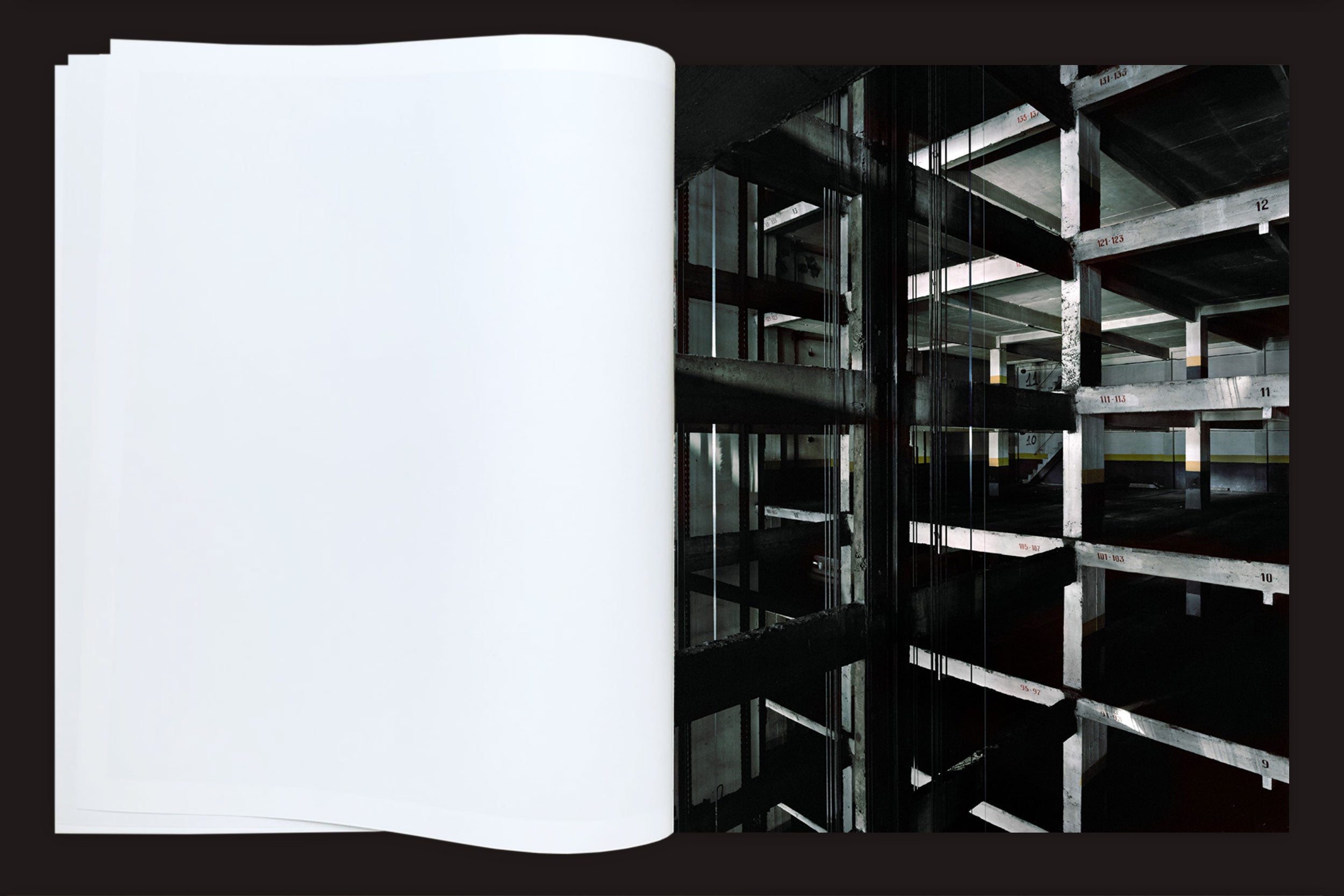 Em Felipe Russo: Garagem Automática by Auto-edição, um livro aberto exibe uma página em branco à esquerda e uma fotografia arquitetônica de uma estrutura de estacionamento de concreto escuro e inacabado com vigas expostas e pisos numerados à direita.