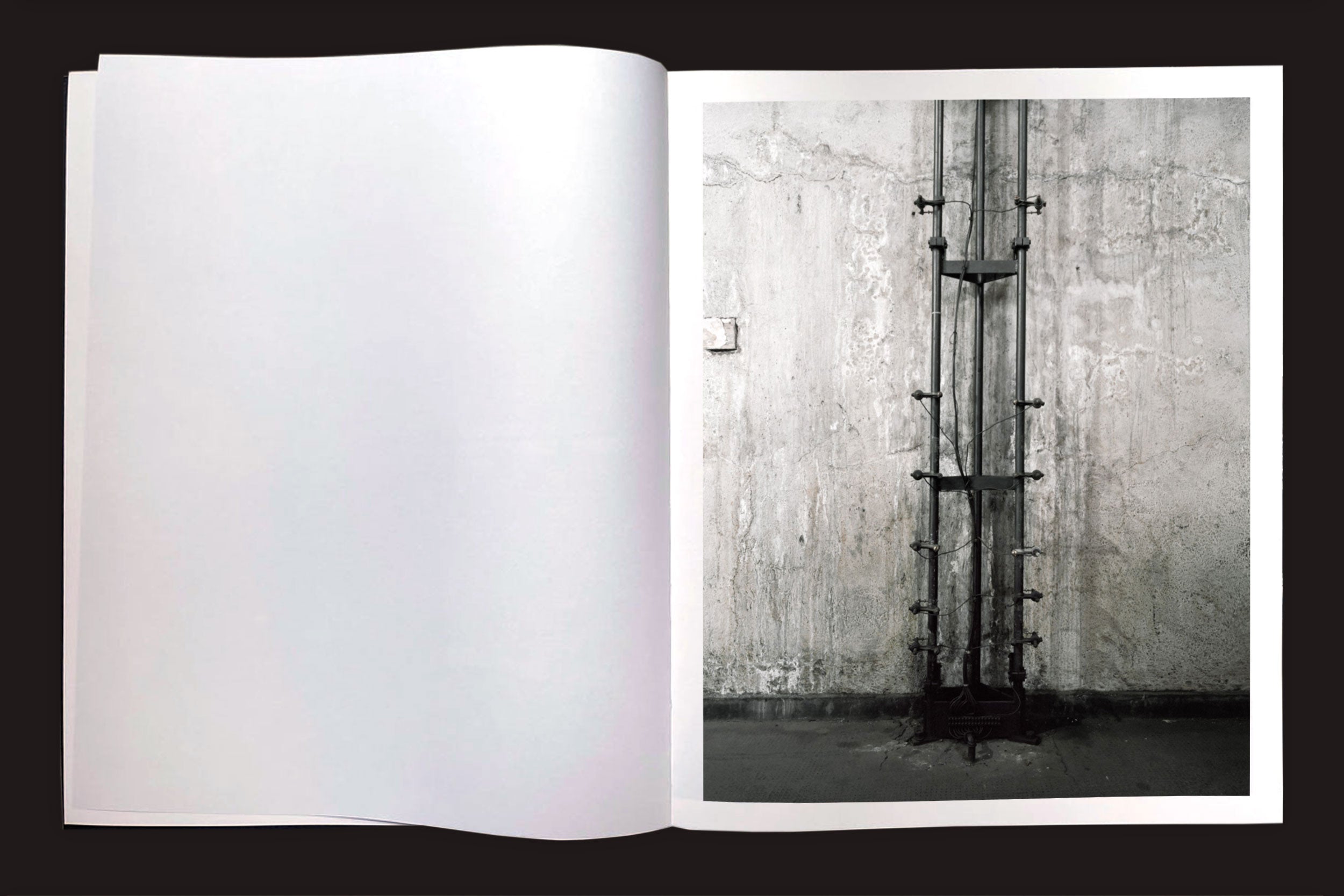 Em "Felipe Russo: Garagem Automática", da Auto-edição, um livro aberto em uma superfície escura mostra uma foto arquitetônica em preto e branco de tubos de metal em uma parede texturizada, com a página esquerda em branco.
