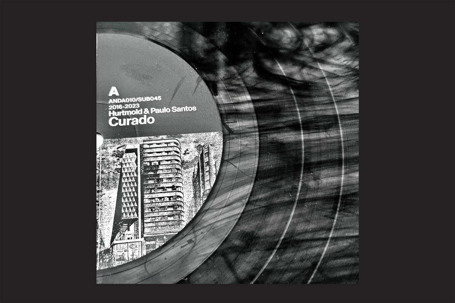 Foto em preto-e-branco do vinil "Hurtmold, Paulo Santos: Curado", vinil da Nada Nada Discos. O selo mostra uma paisagem urbana e informações de catálogo, destacando a música instrumental, com sulcos visíveis e reflexos no disco.