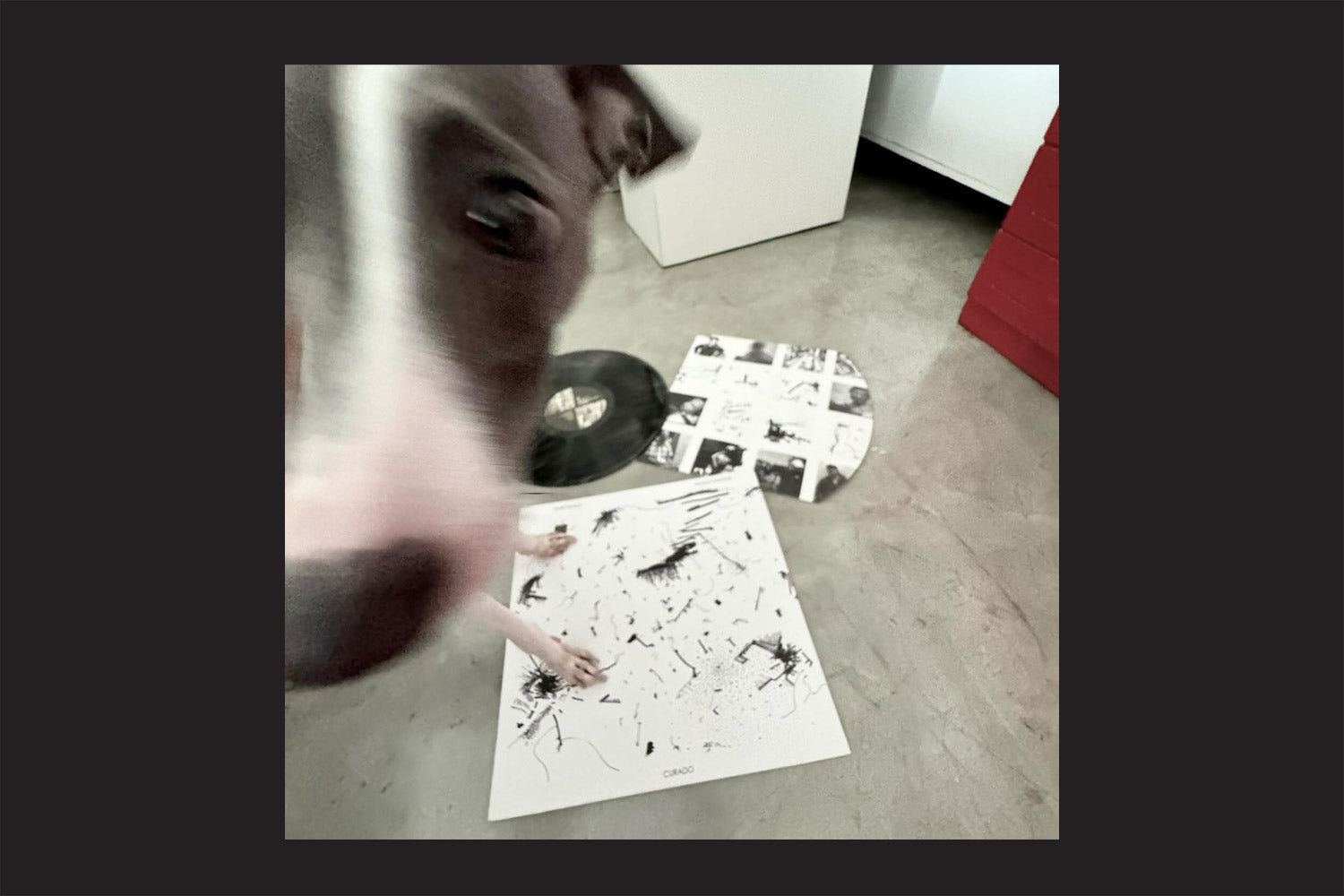 Um cachorro com uma faixa branca no rosto olha para a câmera, parado sobre um disco de vinil da Nada Nada Discos "Hurtmold, Paulo Santos: Curado" e uma obra de arte abstrata em preto e branco em um piso de concreto.