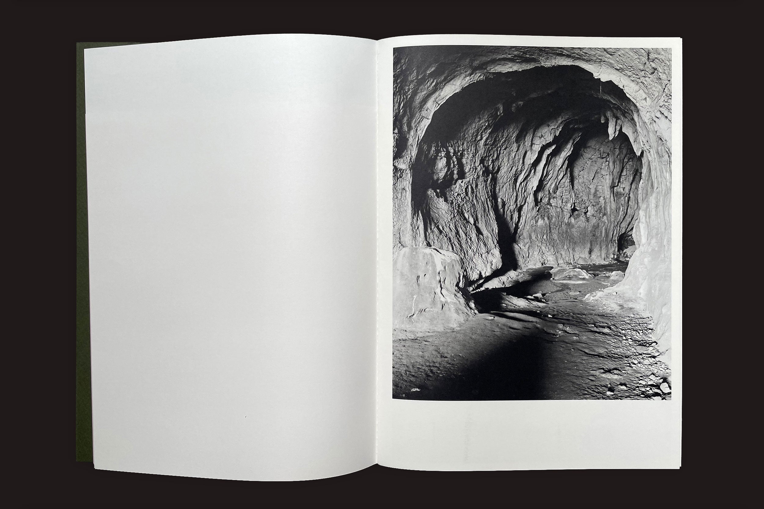 Uma cópia aberta de Felipe Russo: Lugar Dito by Auto-edição revela uma foto em preto e branco da entrada de uma caverna rochosa; paredes texturizadas e pedras espalhadas contrastam fortemente com a página esquerda em branco do livro.